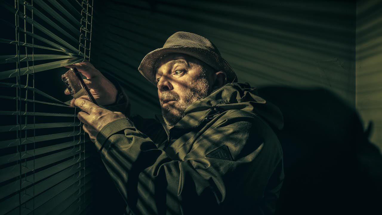 Home A man in a hat suspiciously peering through blinds, holding a phone in shadowy light.